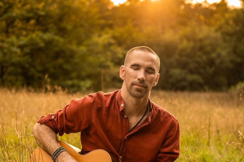Jorn Ayan seated in tall grass playing guitar at golden hour